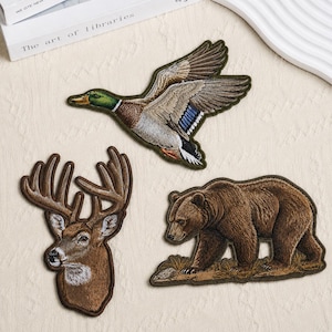 Könnte beinhalten: Drei gestickte Aufnäher mit Wildtieren: eine fliegende Stockente mit grünem, braunem und blauem Gefieder, ein brauner Hirschkopf mit großem Geweih und ein brauner Bär, der auf Gras läuft. Jeder Aufnäher hat einen dunkelbraunen Rand.