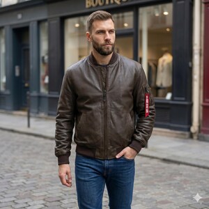 May include: A dark brown leather bomber jacket with a ribbed collar, cuffs, and hem. The jacket features a central zip closure and a small utility pocket on the left sleeve with a red fabric tag attached to the zip pull, worn with blue jeans.