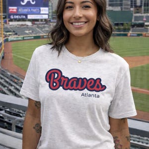 Op de afbeelding: Een lichtgrijs t-shirt met het woord "Braves" in rode letters met een marineblauwe omlijning, en "Atlanta" eronder. Het t-shirt wordt gedragen door een persoon in een honkbalstadion.