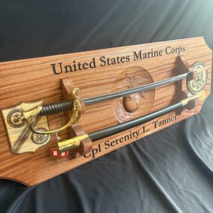 Könnte beinhalten: Eine Holzvitrine mit Schwert und Scheide, mit goldenen Akzenten. Die Vitrine ist mit "United States Marine Corps" und "Cpl Serenity L. Tanner" graviert. Das Marine Corps-Emblem ist ebenfalls vorhanden. Das Schwert hat einen goldenen Knauf und einen schwarzen Griff.