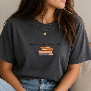 Può includere: T-shirt grigio scuro con la scritta "Books First, Everything else later." Sotto, una pila di libri arancioni, marroni e viola. La maglietta è in stile casual.