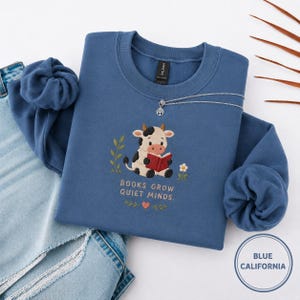 Puede incluir: Una sudadera azul con un gráfico de una vaca leyendo un libro, con el texto "BOOKS GROW QUIET MINDS". Un collar plateado con un colgante de árbol está en la sudadera. Las palabras "BLUE CALIFORNIA" están en un círculo en la parte inferior derecha.