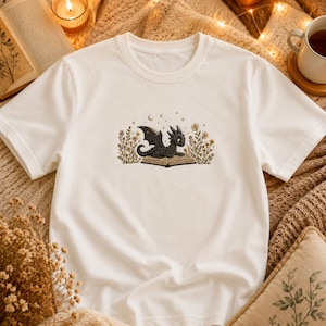 May include: White t-shirt with an embroidered design of a black dragon on an open book, surrounded by flowers and stars. The dragon has detailed wings and a textured appearance. The shirt has a classic crew neck.