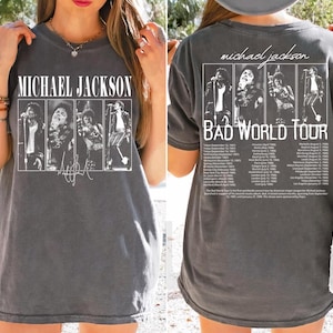 May include: Dark gray t-shirt featuring a graphic of Michael Jackson with the text "MICHAEL JACKSON" above and his signature below. The back of the shirt displays "michael jackson BAD WORLD TOUR" with tour dates and images.