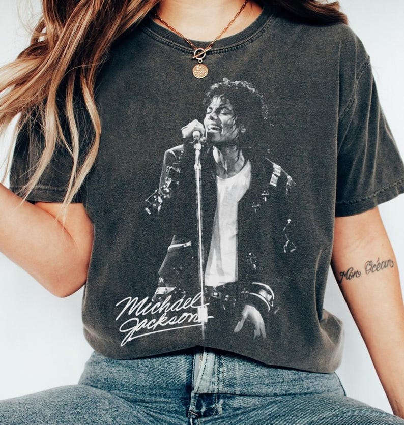 May include: Dark gray t-shirt featuring a black and white image of Michael Jackson performing on stage. The shirt has the text "Michael Jackson" in a white script font. The shirt is worn with blue jeans.