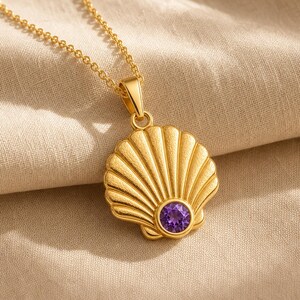 May include: A gold-toned necklace featuring a textured seashell pendant. The pendant is shaped like a fan-style scallop shell with a single, round, faceted purple gemstone set at the base. The pendant hangs from a delicate, matching gold-toned chain.