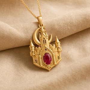 May include: A gold pendant necklace featuring a detailed castle structure with a crescent moon and a star at the top. The center of the castle is adorned with a single, oval-shaped, deep red gemstone. The pendant hangs from a delicate gold chain.