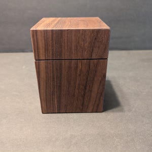 May include: A square wooden box with a dark brown finish and visible natural wood grain patterns. The box features a lid that sits flush with the base, creating a seamless appearance. The construction is simple and geometric, showcasing the texture of the wood.
