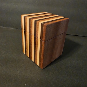 May include: A rectangular wooden box featuring a lid and a body constructed from alternating vertical stripes of light, medium, and dark brown wood. The box has a smooth finish and a clean, geometric design with a horizontal seam separating the lid.