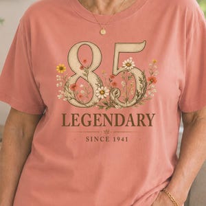 May include: A coral-coloured t-shirt featuring a graphic design with the large number 85 intertwined with colourful wildflowers. Below the number, the text reads LEGENDARY in a bold font, followed by the smaller text SINCE 1941 centred below.
