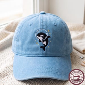 Puede incluir: Una gorra de béisbol azul claro con un diseño bordado de una orca negra y blanca que lleva un casco espacial transparente, rodeada de pequeñas estrellas amarillas de cuatro puntas. La gorra tiene una corona estructurada y visera curva.