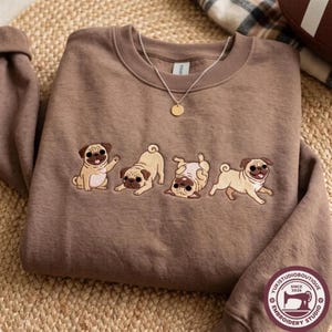 Könnte beinhalten: Ein braunes Sweatshirt mit Rundhalsausschnitt und einer Stickerei von vier beigefarbenen Möpsen in verschiedenen verspielten Posen. Die Möpse sitzen, strecken sich, rollen auf dem Rücken und rennen. Eine feine Goldkette liegt am Kragen.