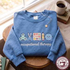 May include: A blue crewneck sweatshirt featuring an embroidered graphic of five smiling objects: a fidget spinner, scissors, a shape sorter, a pencil, and a ball of yarn. Below the characters, the text reads occupational therapy in white script.