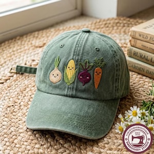 May include: A sage green baseball cap featuring embroidered cartoon vegetables including an onion, a corn cob, a beet, and a carrot, each with a smiling face. The cap has a curved brim and a fabric strap with a metal buckle on the back.