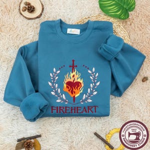 May include: A teal crewneck sweatshirt featuring an embroidered design of a red heart engulfed in yellow and orange flames, pierced by a sword, and framed by white laurel branches. The word FIREHEART is embroidered in red below the graphic.