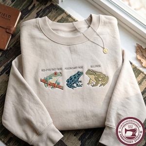 May include: A beige crewneck sweatshirt featuring embroidered illustrations of three frogs with the text labels Red-Eyed Tree Frog, Poison Dart Frog, and Bullfrog. The design includes a green and orange tree frog, a blue spotted frog, and a green bullfrog.