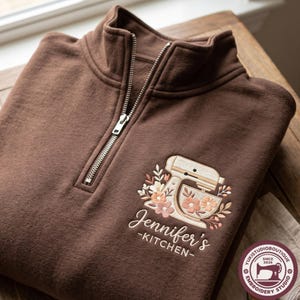 Könnte beinhalten: Ein dunkelbraunes Sweatshirt mit kurzem Reißverschluss und einer Stickerei, die einen Standmixer umgeben von Blumen zeigt. Unter dem Mixer steht der Text "Jennifer's KITCHEN" in weißer Schreib- und Blockschrift. Der Kragen hat einen Reißverschluss.