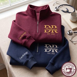 May include: A maroon and a navy blue quarter-zip pullover sweatshirt. The maroon sweatshirt features gold embroidery reading DR Anthony EST. 2026 and Cardiology on the sleeve. The navy sweatshirt features white embroidery reading DR Anthony EST. 2026 and Cardiology.