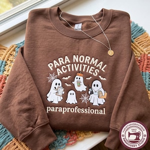 Peut inclure: Un sweat-shirt marron à col rond orné d'un motif représentant trois fantômes blancs avec le texte "PARA NORMAL ACTIVITIES" et "paraprofessional". Les fantômes tiennent un presse-papier, un café, un talkie-walkie et un dossier, entourés de toiles d'araignée.