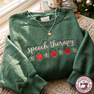 May include: A forest green crewneck sweatshirt featuring the embroidered text "speech therapy" in white cursive script, accented by a row of three red strawberries and four white flowers. A thin gold chain necklace rests on the neckline.