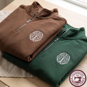 Peut inclure: Un sweat-shirt marron et un sweat-shirt vert foncé à fermeture éclair quart, chacun arborant une broderie blanche en forme de cerveau sur la poitrine. Ils possèdent un col montant avec une fermeture éclair argentée. Un logo circulaire est présent.