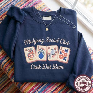 May include: A navy blue crewneck sweatshirt featuring an embroidered graphic of four mahjong tiles with a dragon, a vase of flowers, a bird on a branch, and a floral design. The text reads Mahjong Social Club and Crak Dot Bam in light pink.