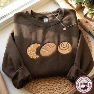 May include: A dark brown crewneck sweatshirt featuring an embroidered design of a croissant, a loaf of bread, and a cinnamon roll in shades of tan and gold. A delicate gold-tone chain necklace rests on the upper right side of the garment.