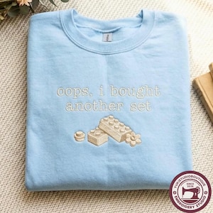Puede incluir: Una sudadera de cuello redondo en color azul claro con bordado en beige que dice "oops, i bought another set" sobre un gráfico de dos bloques de construcción de plástico entrelazados y una pequeña pieza con forma de flor.