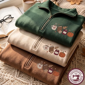 May include: Three quarter-zip sweatshirts in dark green, beige, and brown, each featuring an embroidered design of a mug with a heart, a closed book, a candle, and a pumpkin, accented with small leaves and star shapes on the upper left chest.