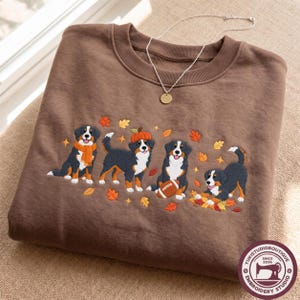 Puede incluir: Una sudadera marrón de cuello redondo con un diseño bordado de cuatro perros de montaña de Berna. Los perros aparecen con elementos otoñales, como hojas naranjas, una calabaza y un balón de fútbol americano. Un collar de plata descansa sobre el cuello.