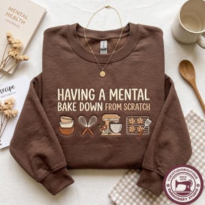 May include: A dark brown crewneck sweatshirt featuring the text "HAVING A MENTAL BAKE DOWN FROM SCRATCH" in cream lettering. Below the text are graphic illustrations of stacked bowls, a whisk, a stand mixer, and a tray of cookies with a spatula.