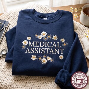 May include: A navy blue crewneck sweatshirt featuring the white embroidered text MEDICAL ASSISTANT centred on the chest. The text is surrounded by a circular arrangement of embroidered white and yellow daisy flowers with green stems.