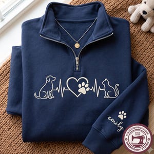 May include: A navy blue quarter-zip sweatshirt featuring a white embroidered heartbeat line graphic with a dog silhouette, a heart with a paw print, and a cat silhouette. The sleeve is embroidered with the name emily and a small paw print.