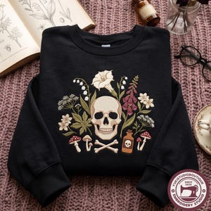 Peut inclure: Un sweat-shirt noir à col rond orné d'un motif central représentant un crâne humain entouré d'illustrations botaniques, dont des fleurs blanches, des digitales violettes, des champignons rouges, une fiole brune et des os croisés.