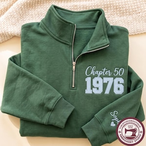 May include: A dark green quarter-zip sweatshirt featuring embroidered light blue text that reads Chapter 50 and 1976 on the chest. The left sleeve includes the embroidered name Sophie with a small heart icon near the ribbed cuff.