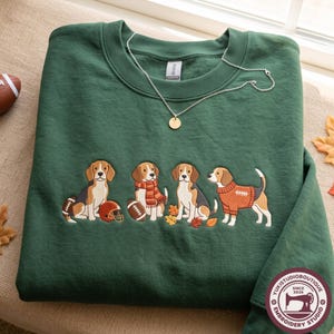 Op de afbeelding: Een bosgroene sweater met ronde hals en een geborduurd ontwerp van vier beagle-honden. De honden zijn afgebeeld met footballs, een footballhelm, een oranje sjaal en een oranje trui, omringd door kleine herfstbladeren.