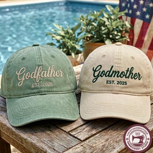 Peut inclure: Deux casquettes de baseball, une vert sauge et une beige, avec du texte brodé. La casquette verte affiche 'Godfather' avec 'EST. 2026' en dessous. La casquette beige affiche 'Godmother' avec 'EST. 2025' en dessous. Visières courbées.
