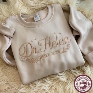 May include: A beige crewneck sweatshirt featuring tan embroidery that reads Dr. Helen, PHD 2026. The design includes small star accents and two leafy branch motifs flanking the text. The garment has a ribbed collar, cuffs, and hemline.
