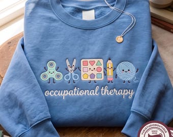 May include: A light blue crewneck sweatshirt featuring embroidered cartoon characters of a fidget spinner, scissors, a shape sorter, a pencil, and a ball of yarn, with the white text "occupational therapy" stitched below the figures.