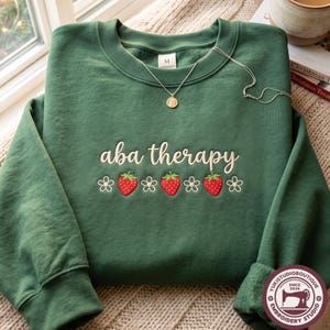 Puede incluir: Una sudadera de cuello redondo color verde bosque con el texto bordado "aba therapy" en letra cursiva blanca, centrado sobre una fila de tres fresas rojas y cuatro flores blancas. Un collar de cadena plateada con un dije redondo descansa encima.