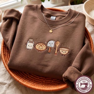 May include: A chocolate brown crewneck sweatshirt featuring an embroidered design of five smiling kitchen items: a glass jar, a round loaf of bread, a metal dough scraper, a wooden spoon, and a mixing bowl, accented with small embroidered stars.