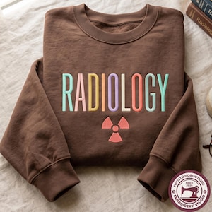 May include: A brown crewneck sweatshirt featuring the word RADIOLOGY embroidered in multi-coloured pastel letters across the chest. Below the text is a coral-coloured radiation symbol. The sweatshirt has long sleeves and a ribbed collar.