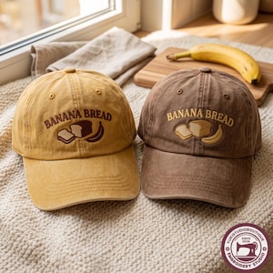 May include: Two baseball caps, one mustard yellow and one brown, each featuring embroidered text that reads BANANA BREAD above a graphic of a loaf of bread, a slice of bread, and a banana. The caps have a curved brim and stitched eyelets.