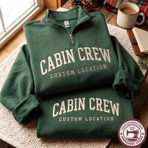 May include: A dark green quarter-zip sweatshirt featuring the text CABIN CREW and CUSTOM LOCATION in cream-colored lettering. A matching dark green garment is folded underneath, also displaying the text CABIN CREW and CUSTOM LOCATION in cream.