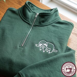 May include: A dark green quarter-zip sweatshirt featuring a white embroidered sloth resting on a branch. The garment has a high collar with a silver-toned metal zip closure. A circular logo with the text YUKISTUDIOBOUTIQUE SINCE 2026 is visible.