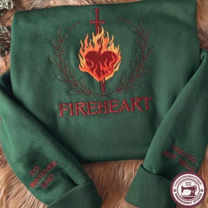 May include: A forest green sweatshirt featuring an embroidered design of a red heart surrounded by orange and yellow flames, pierced by a sword, and framed by laurel branches. The text FIREHEART is below, with TO WHATEVER END and YOU DO NOT YIELD on sleeves.