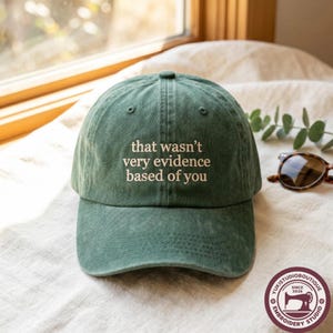 May include: A forest green baseball cap with a distressed, washed-out texture. The front of the cap features the embroidered text "that wasn't very evidence based of you" in a light cream colour, centred in a simple, clean, sans-serif font.