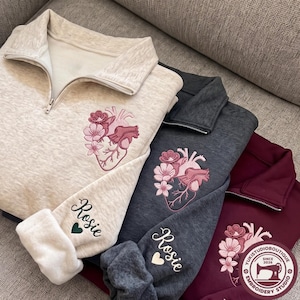 May include: Three quarter-zip sweatshirts in oatmeal, charcoal gray, and maroon. Each features an embroidered anatomical heart with pink flowers on the chest and the name Rosie with a small heart on the sleeve cuff. The fabric has a soft texture.