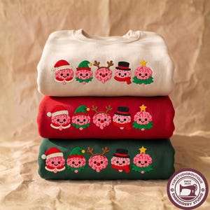 May include: A stack of three crewneck sweatshirts in cream, red, and dark green. Each features an embroidered row of five pink cartoon brains dressed as holiday characters: a Santa, an elf, a reindeer, a snowman, and a Christmas tree.