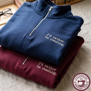 May include: A navy blue and a maroon quarter-zip sweatshirt, each featuring the embroidered white text "I'd rather be reading." The sweatshirts have a high collar with a silver-toned metal zipper closure and a soft, solid-colored fabric construction.